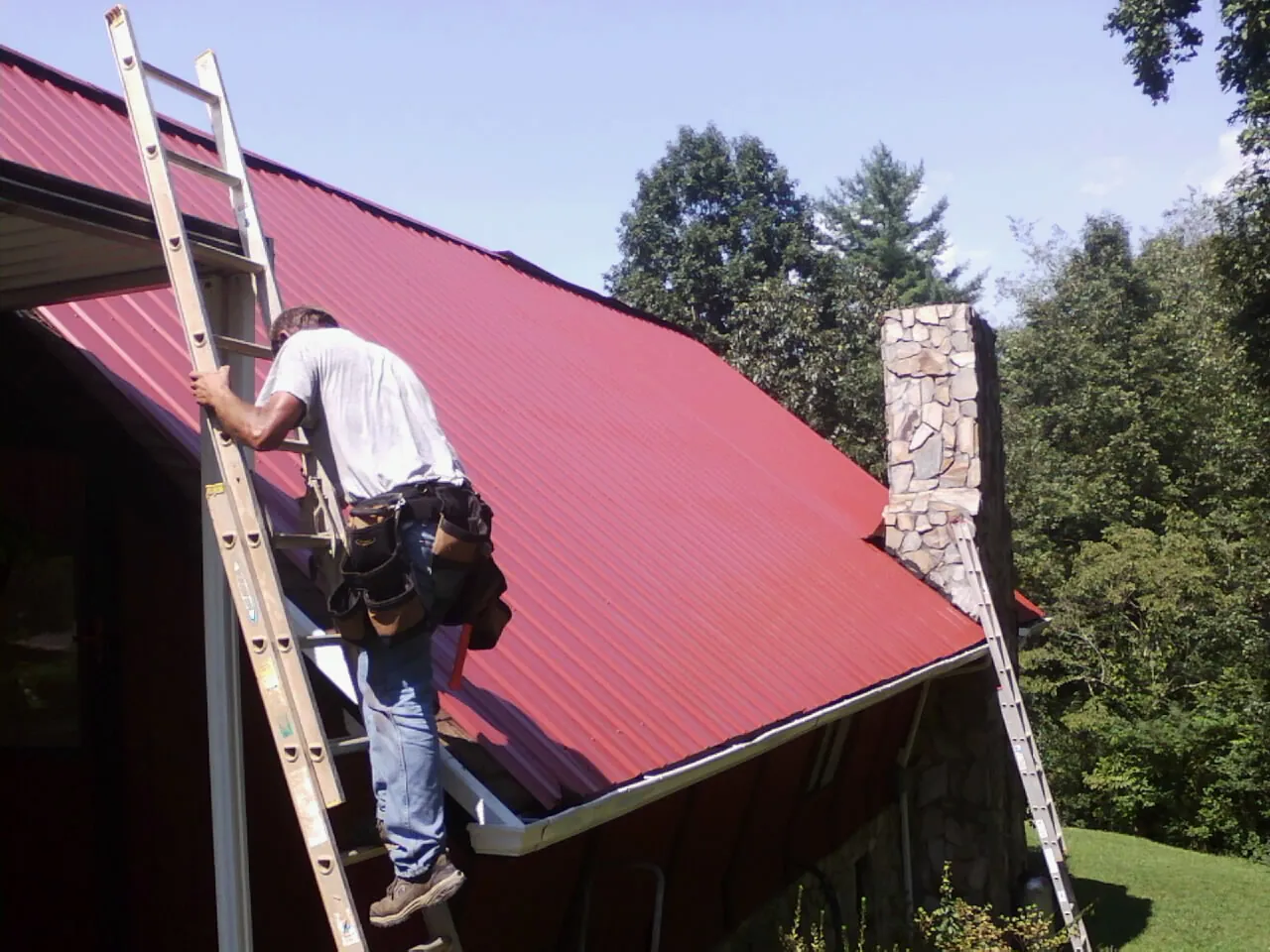 Roofer on a ladder beside a red metal roof and stone chimney — Soffit & Fascia Repair by Roofing Brothers in Dougherty